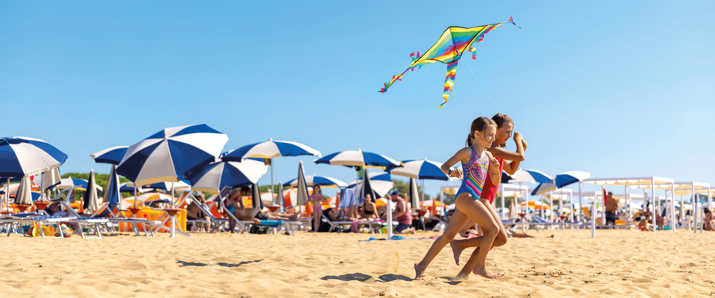 Due bambine corrono sulla spiaggia di Bibione sotto un aquilone colorato